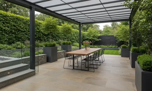 Modern outdoor dining area with a long wooden table, black chairs, overhead canopy, stone steps, and lush greenery surrounding a minimalist garden with glass railing