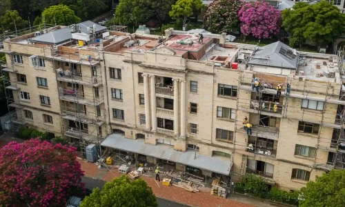 View of a sandstone building with classical columns and balcony, workers on the roof, and construction scaffolding below View of a sandstone building with classical columns and balcony, workers on the roof, and construction scaffolding below